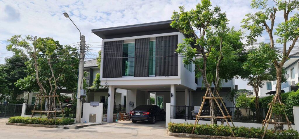 บ้านโมเดิร์นสีขาวมีแผงระแนงแนวตั้งสีเข้มหน้ากระจกชั้นบนและต้นไม้ด้านหน้า