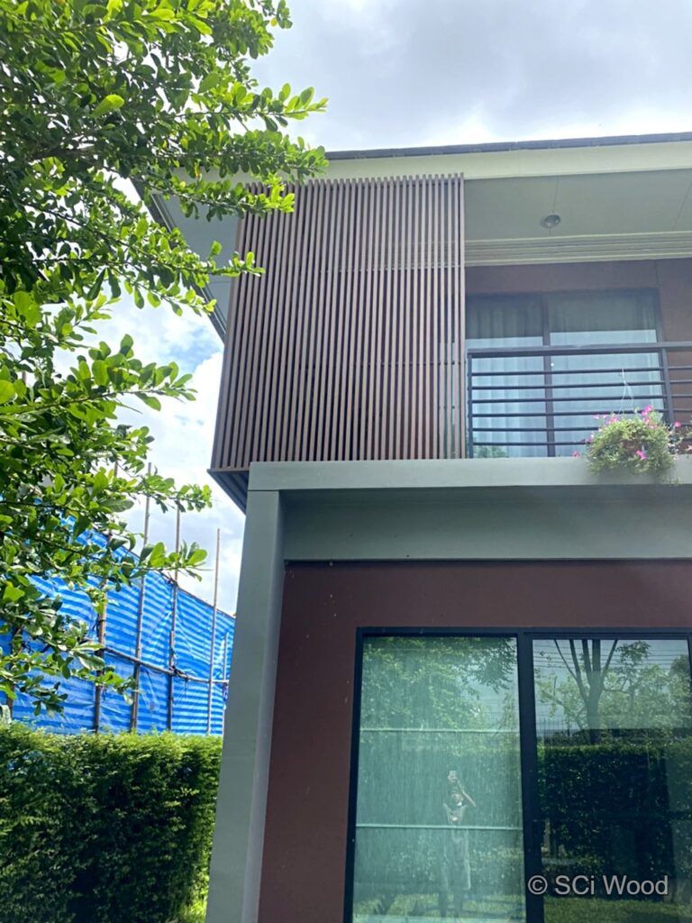 แผงระแนงแนวตั้งข้างระเบียงชั้นสองของบ้าน มีต้นไม้สีเขียวด้านหน้า